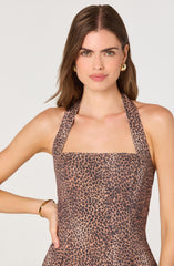 A woman is wearing a sleeveless dress with a square neckline and wide straps. The dress features a leopard print pattern with black spots on a brown background. She accessorized with gold earrings and a gold bracelet.