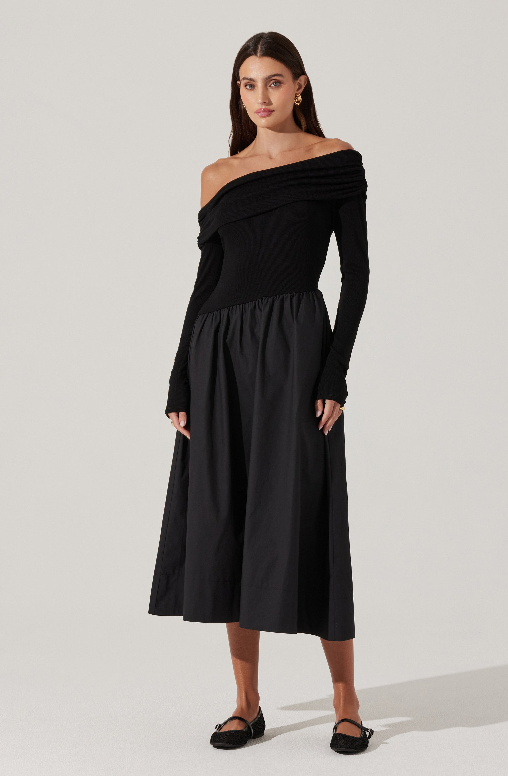 A woman is wearing an off-the-shoulder black dress with a fitted bodice and long sleeves. The dress features a gathered, draped fabric across one shoulder and a full, gathered skirt that reaches mid-calf length. She is also wearing black ballet flats and gold earrings.