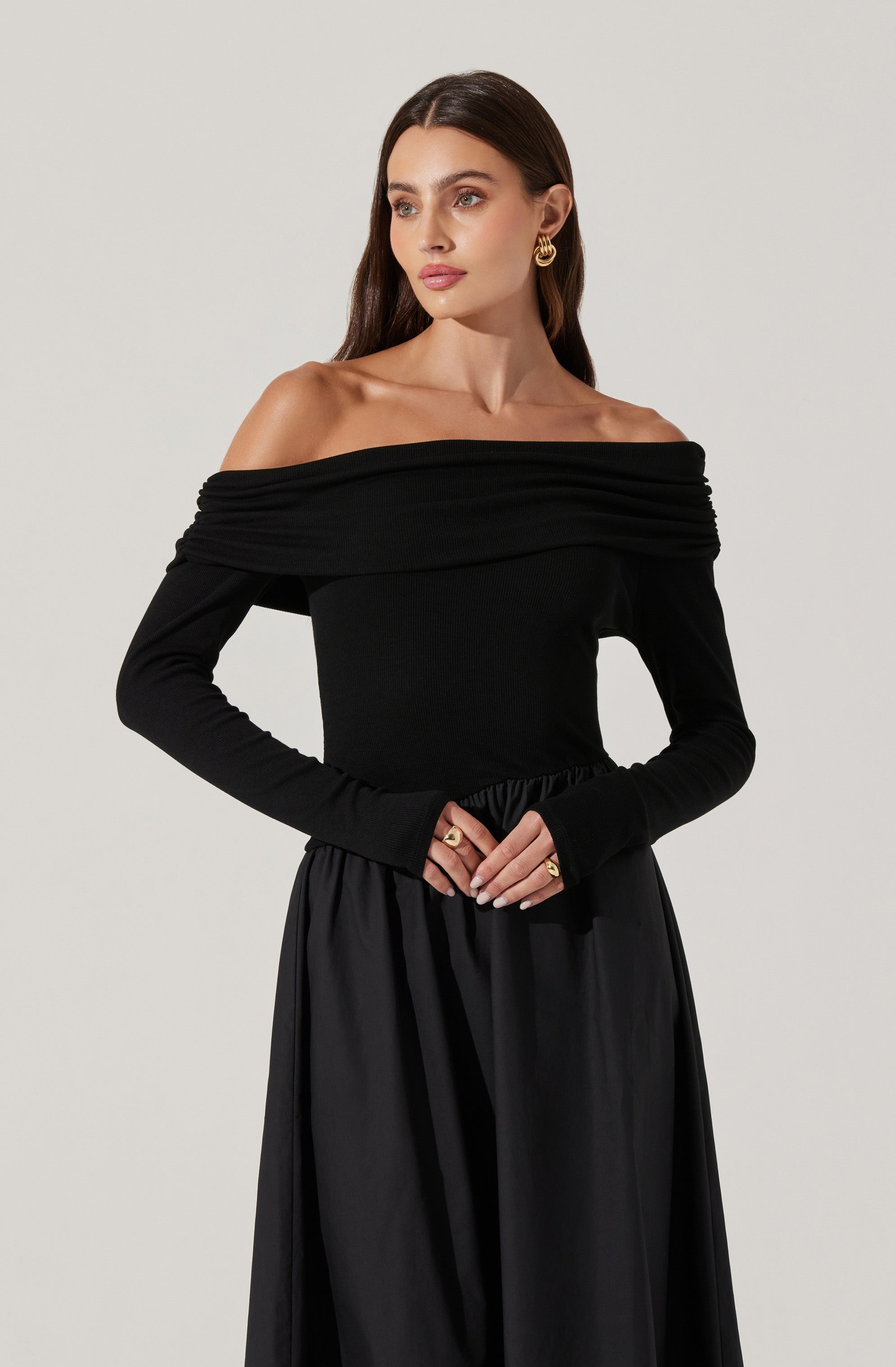 The woman is wearing an off-the-shoulder black top with a wide, draped neckline that extends around her shoulders and arms. The top has long sleeves that fit snugly, and the fabric appears soft and slightly stretchy. She is also dressed in a black, high-waisted skirt with a loose, flowing silhouette made from a lightweight material.
