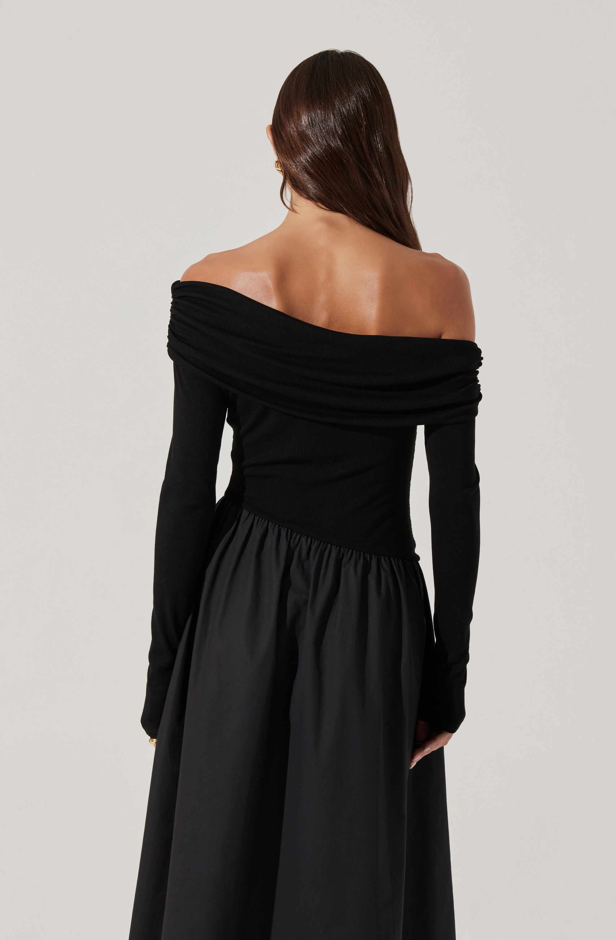 A woman is wearing an off-the-shoulder black dress with a fitted bodice and long sleeves. The top features a wide, ruched neckline that drapes across her shoulders, exposing her back and shoulders. The skirt is full and gathered, creating a flowing silhouette.