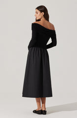 A woman is wearing an off-the-shoulder black dress with long sleeves. The top part of the dress is fitted and gathered at the shoulders, creating a ruched effect. The skirt is high-waisted, full-length, and has a subtle sheen. She is accessorizing with gold earrings and black slip-on shoes.