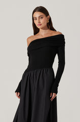 A woman is wearing an off-the-shoulder black dress. The top part of the dress has a ruched design, covering one shoulder and draping across the chest. The skirt is made of a shiny, gathered fabric that creates a full, flowing appearance. She accessorized with large gold hoop earrings and a gold ring.