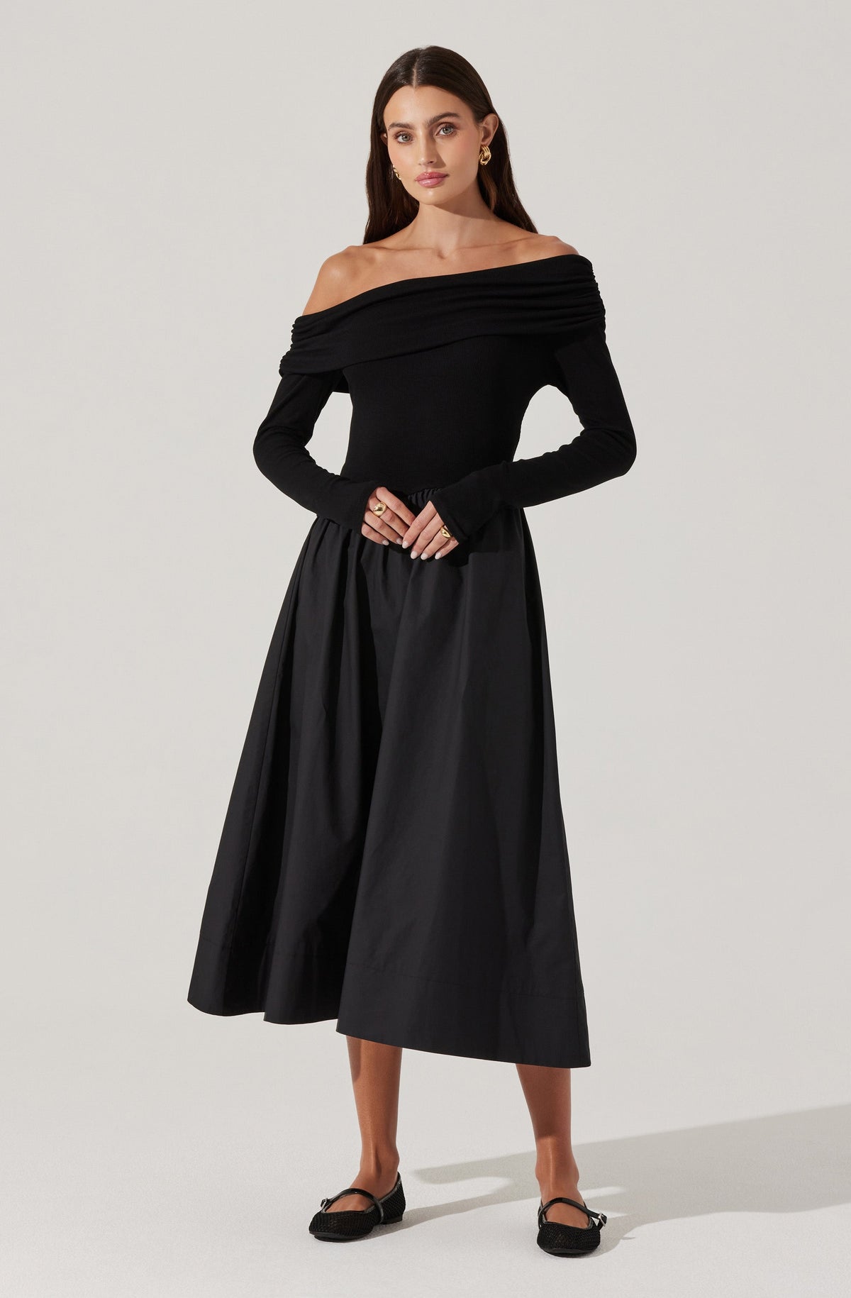A woman is wearing an off-the-shoulder black top with a wide, ruched neckline that drapes across her shoulders. She pairs it with a high-waisted, full-length black skirt that has a slightly shiny, textured fabric. The outfit is accessorized with earrings and rings, and she is wearing black flats.