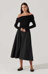 A woman is wearing an off-the-shoulder black top with a wide, ruched neckline that drapes across her shoulders. She pairs it with a high-waisted, full-length black skirt that has a slightly shiny, textured fabric. The outfit is accessorized with earrings and rings, and she is wearing black flats.