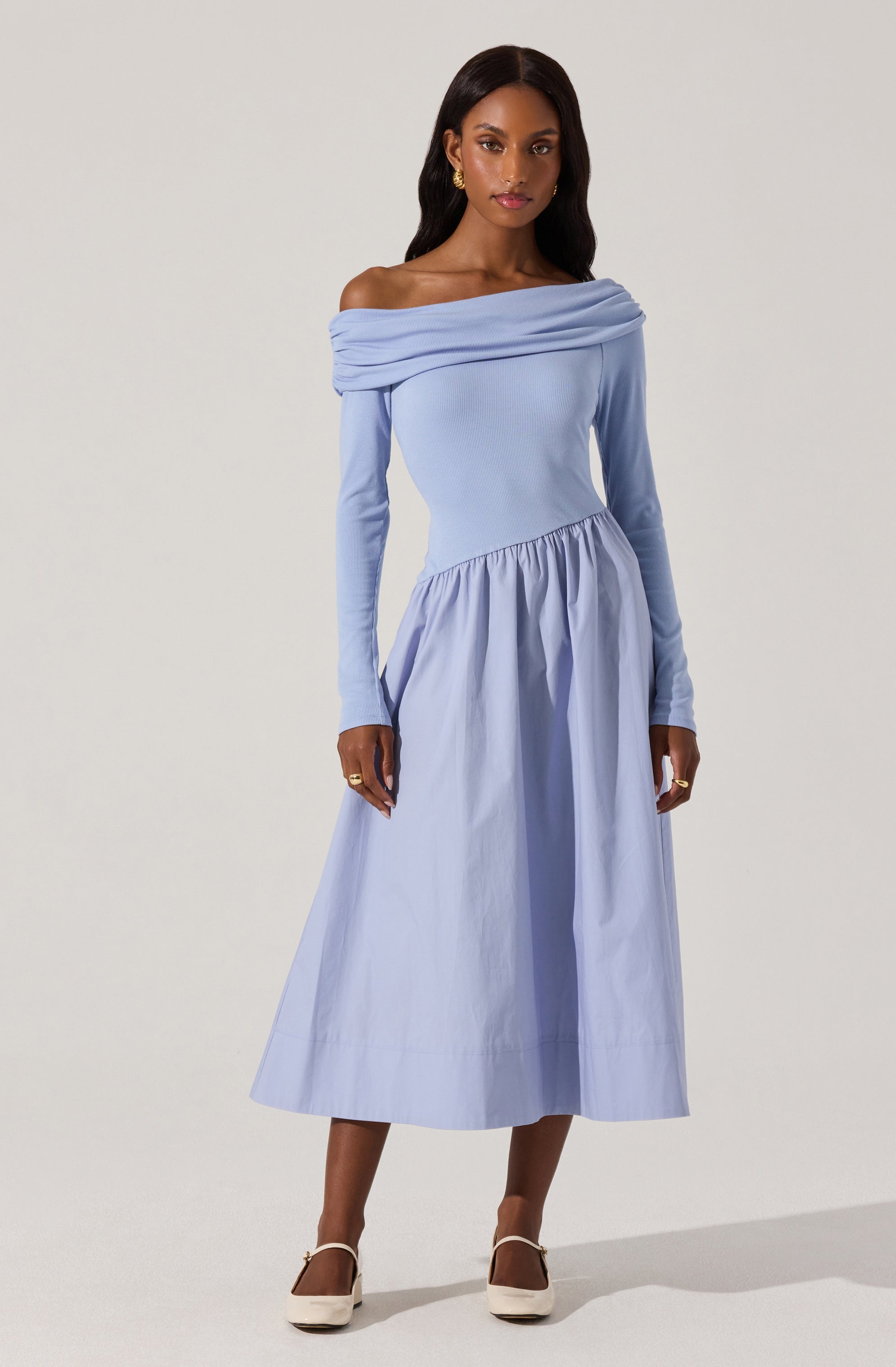 A woman is wearing a light blue, off-the-shoulder dress with long sleeves. The dress features a wide, draped neckline that folds over her shoulders and a fitted bodice that flows into a full, gathered skirt that reaches mid-calf. She pairs the dress with white shoes and gold earrings.