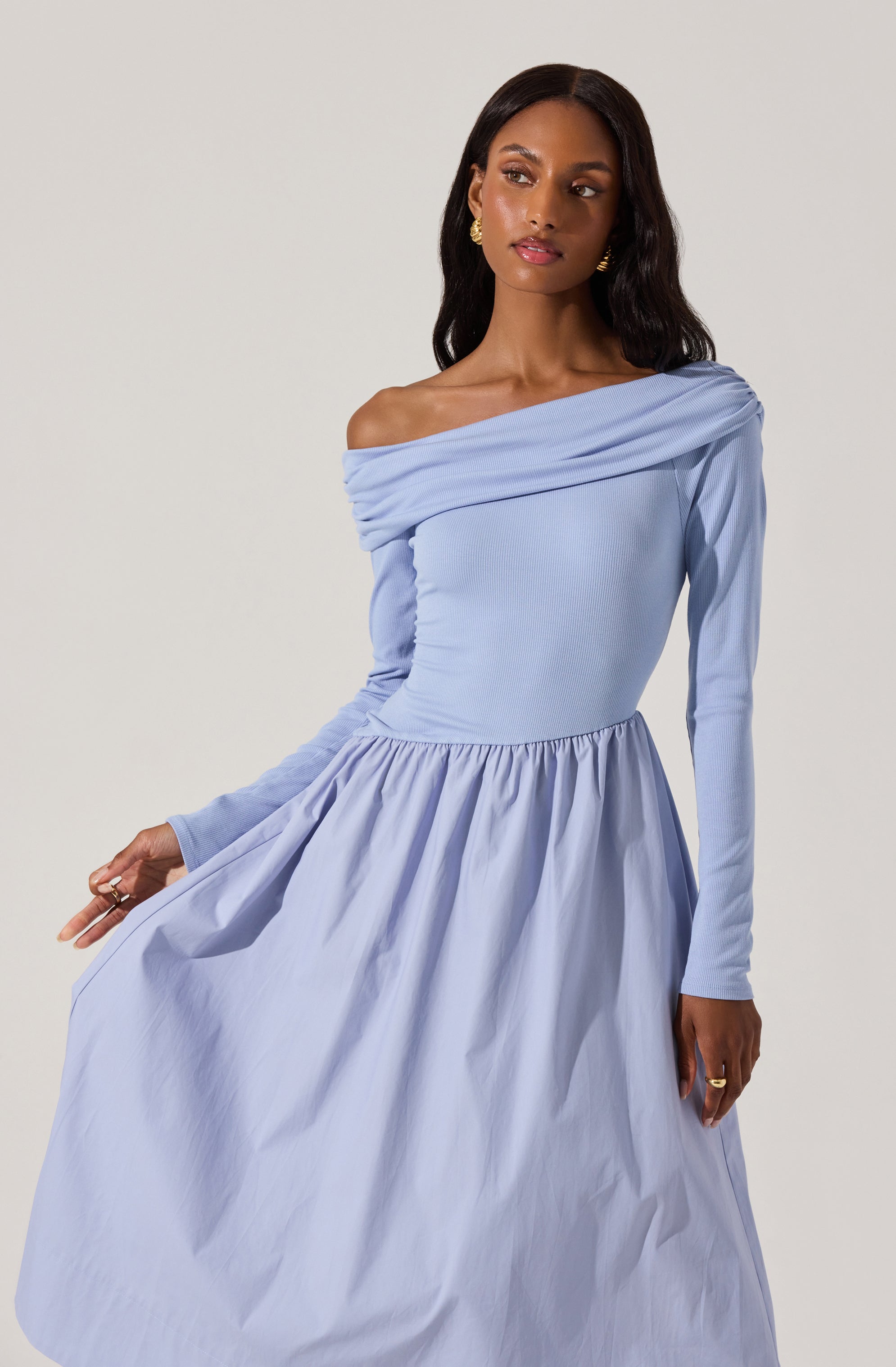 A woman is wearing a light blue off-the-shoulder dress with long sleeves. The dress has a fitted bodice and a full, gathered skirt. The fabric appears to be smooth and slightly shiny, with a draped neckline that exposes her shoulders. She is accessorized with gold earrings and rings.
