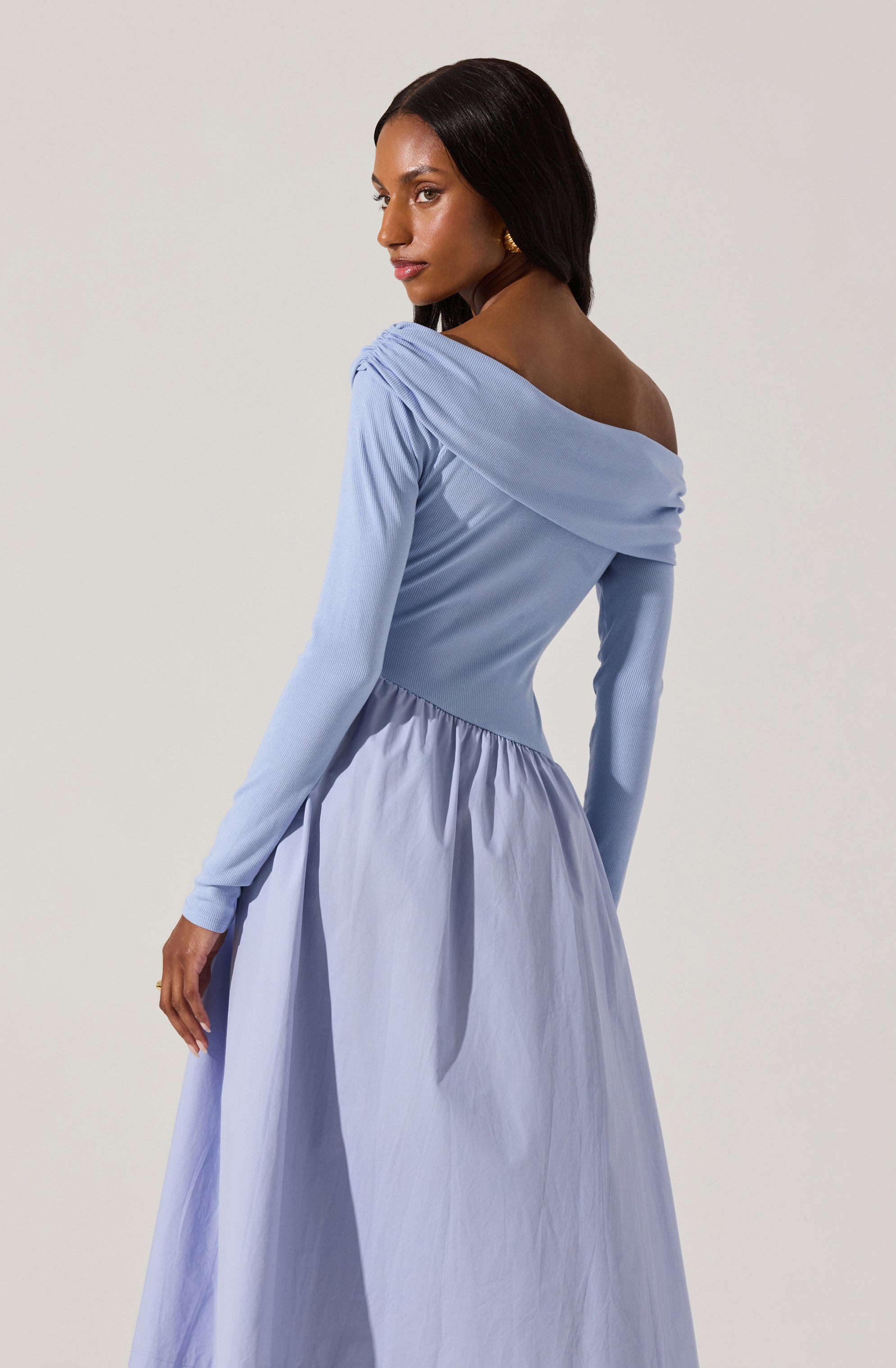 A woman is wearing a long, elegant, light blue dress with long sleeves. The dress features a draped, off-the-shoulder neckline and a fitted bodice that transitions into a full, gathered skirt. The fabric appears smooth and slightly shiny, giving the dress a sophisticated appearance.
