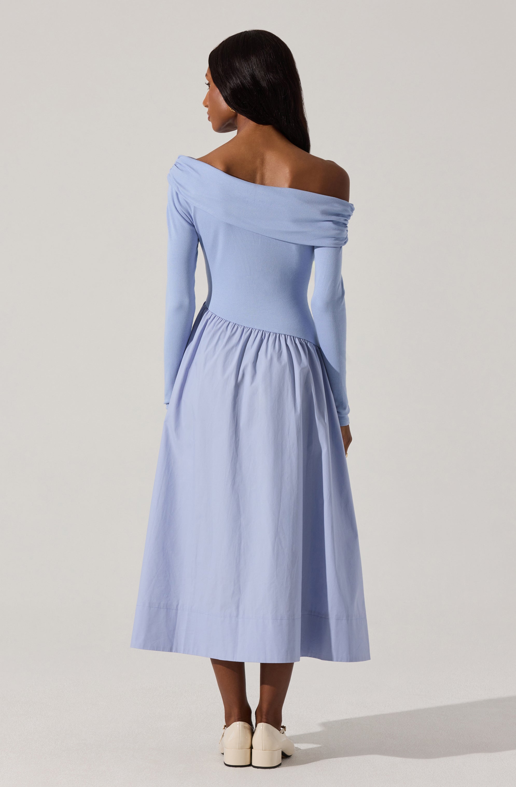 A woman is wearing a long, light blue dress with an off-the-shoulder neckline that features a draped detail across the shoulders. The dress has long fitted sleeves and a gathered, voluminous skirt that falls to her mid-calf. She pairs the dress with white shoes.
