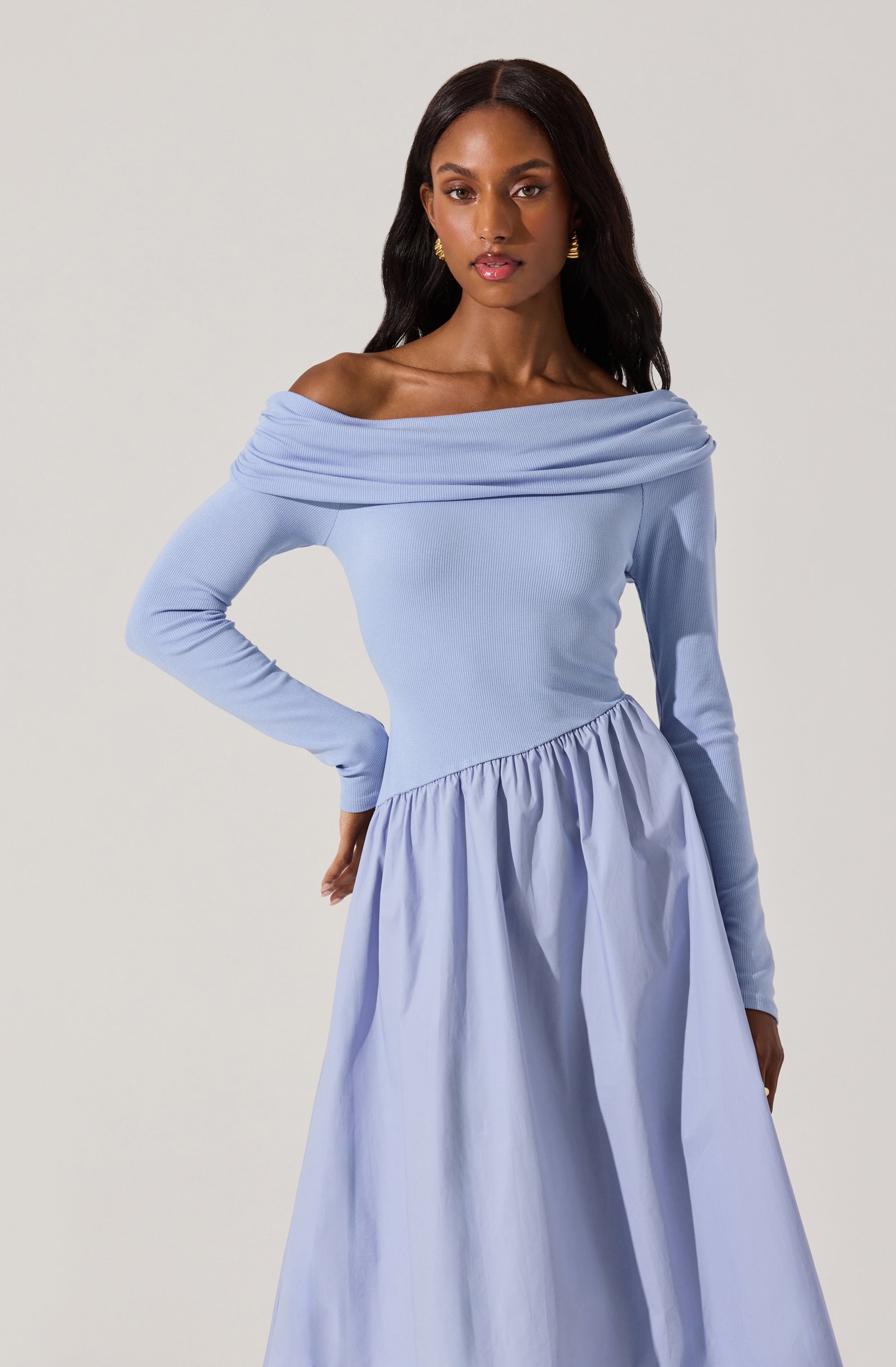 A woman is wearing a light blue, off-the-shoulder dress with long sleeves. The top part of the dress is fitted, and the skirt is gathered and voluminous. The dress has a soft, elegant appearance.