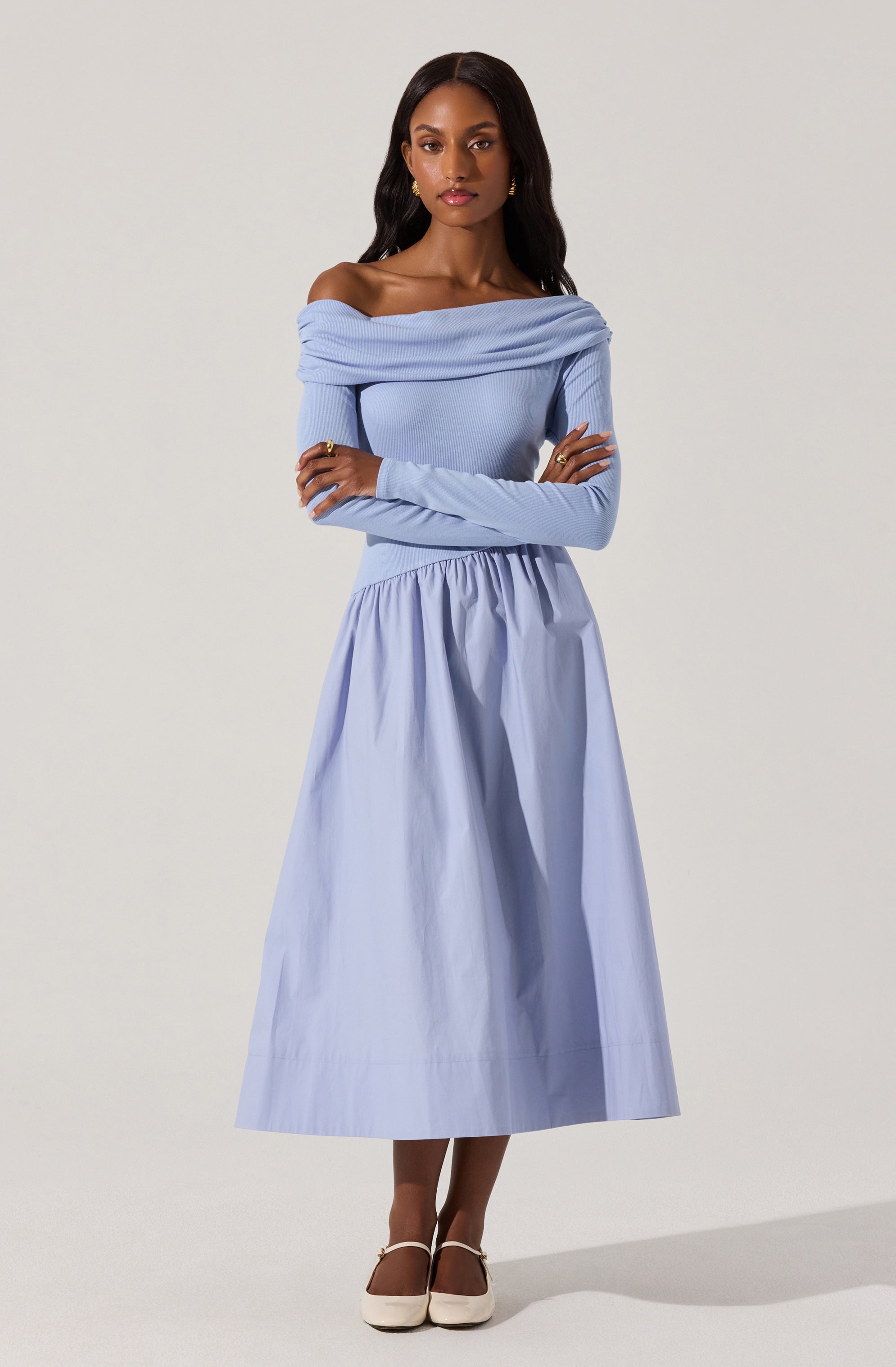 A woman is wearing an off-the-shoulder, long-sleeved, light blue top with a draped neckline, paired with a high-waisted, full-length, light blue skirt with gathered fabric at the waist. She is also wearing white shoes with ankle straps.