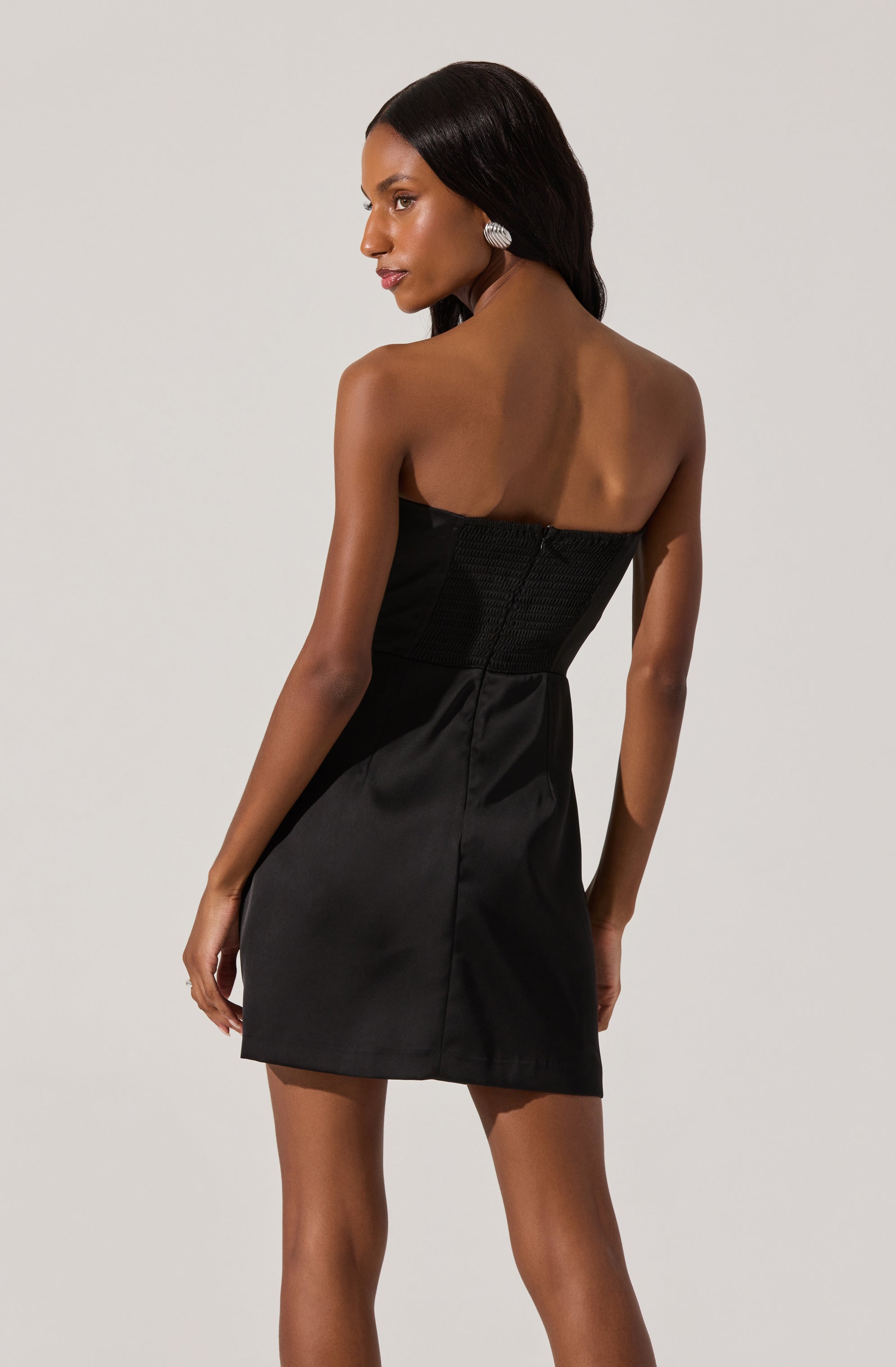 A woman is wearing a strapless black dress that features a fitted, textured bodice and a smooth, slightly shiny skirt. The dress is knee-length, and she accessorizes with large, round, silver earrings.
