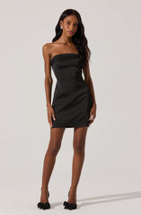 A woman is wearing a strapless, fitted black mini dress with a straight neckline. She has on black high heels with bow details at the toes. The overall look is elegant and sophisticated.