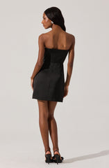 A woman is wearing a strapless black dress with a fitted bodice and a slightly flared skirt. She is also wearing black high heels with ankle straps and sparkly, circular earrings.