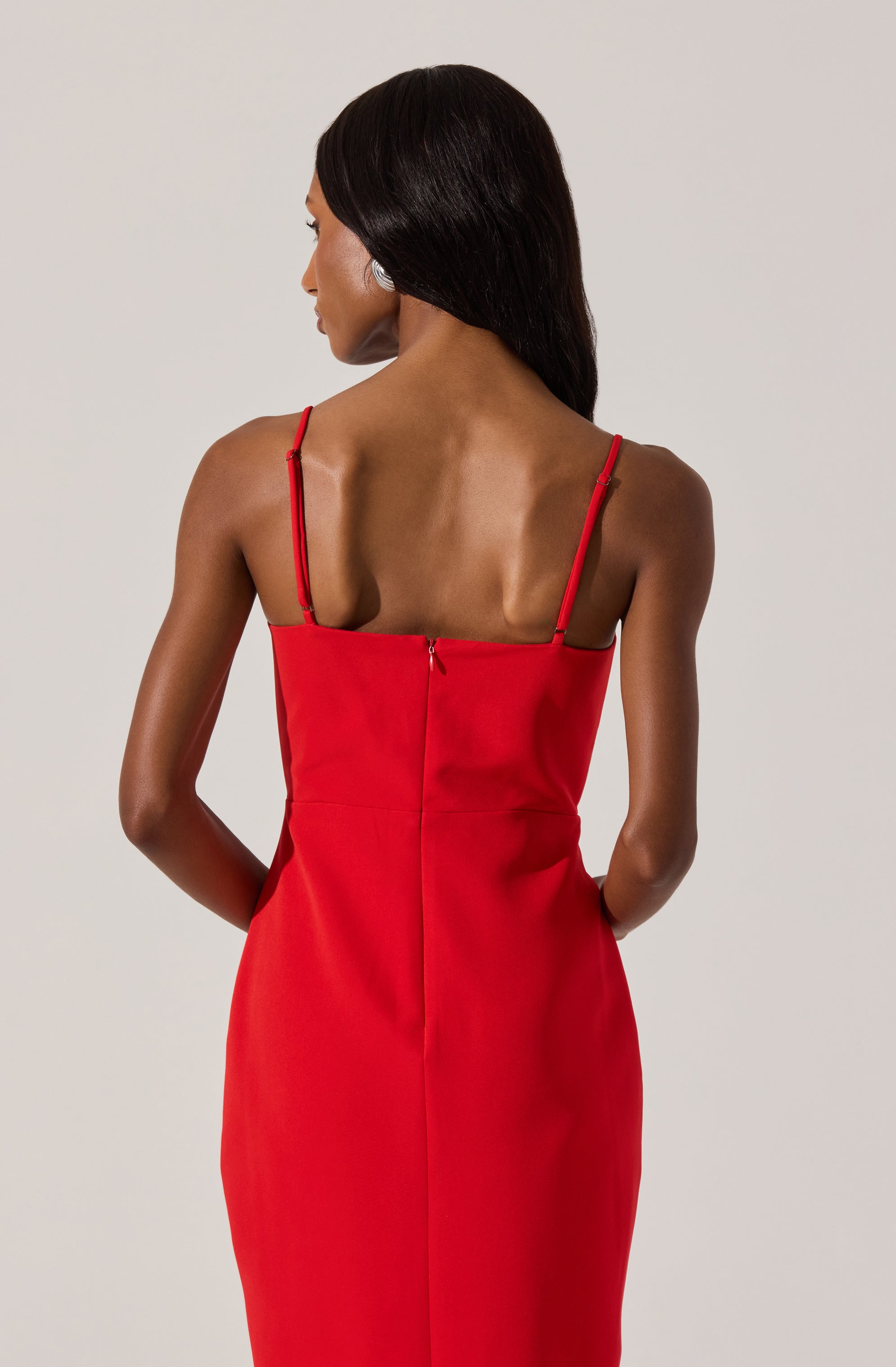 A woman is wearing a form-fitting, sleeveless red dress with thin spaghetti straps. The dress has a zipper at the back and a smooth, elegant fabric.