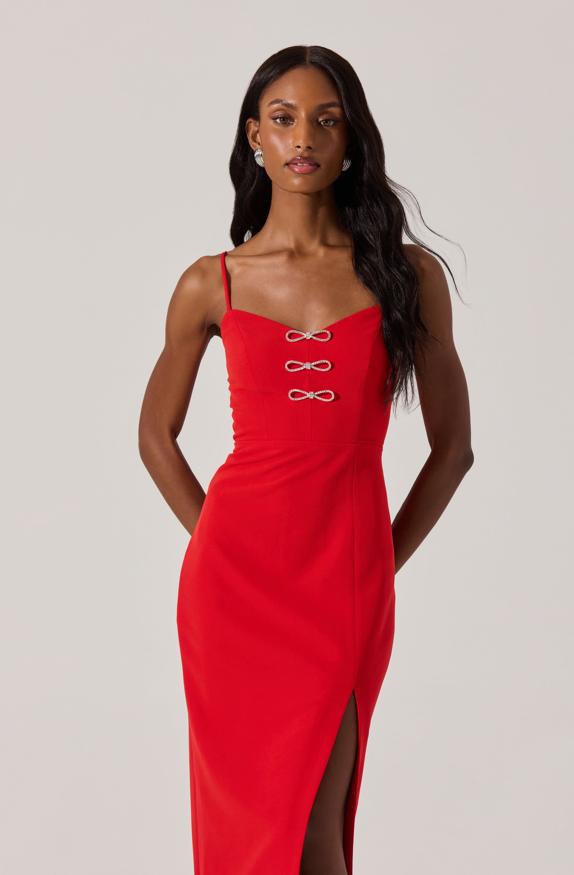 A woman is wearing a fitted, sleeveless red dress with thin spaghetti straps. The dress features a sweetheart neckline and decorative bow-like accents on the front bodice. It has a high slit on the side, revealing part of her leg.