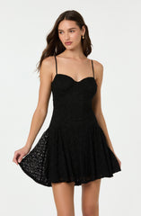 A woman is wearing a black dress with thin spaghetti straps. The top part of the dress is fitted with a sweetheart neckline, and the fabric has a textured, lace-like pattern. The skirt is flared and made of a lightweight, semi-sheer lace material with an intricate pattern, giving it a delicate and feminine appearance.