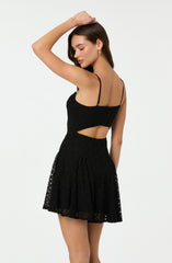 A woman is wearing a black lace dress with thin spaghetti straps. The dress features a fitted bodice with a cutout at the waist and a flared skirt. The lace fabric adds a delicate, textured appearance to the dress.