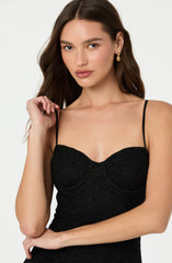 A woman is wearing a black, textured dress with thin spaghetti straps and a fitted bodice. She has gold earrings and her long brown hair is styled in loose waves.