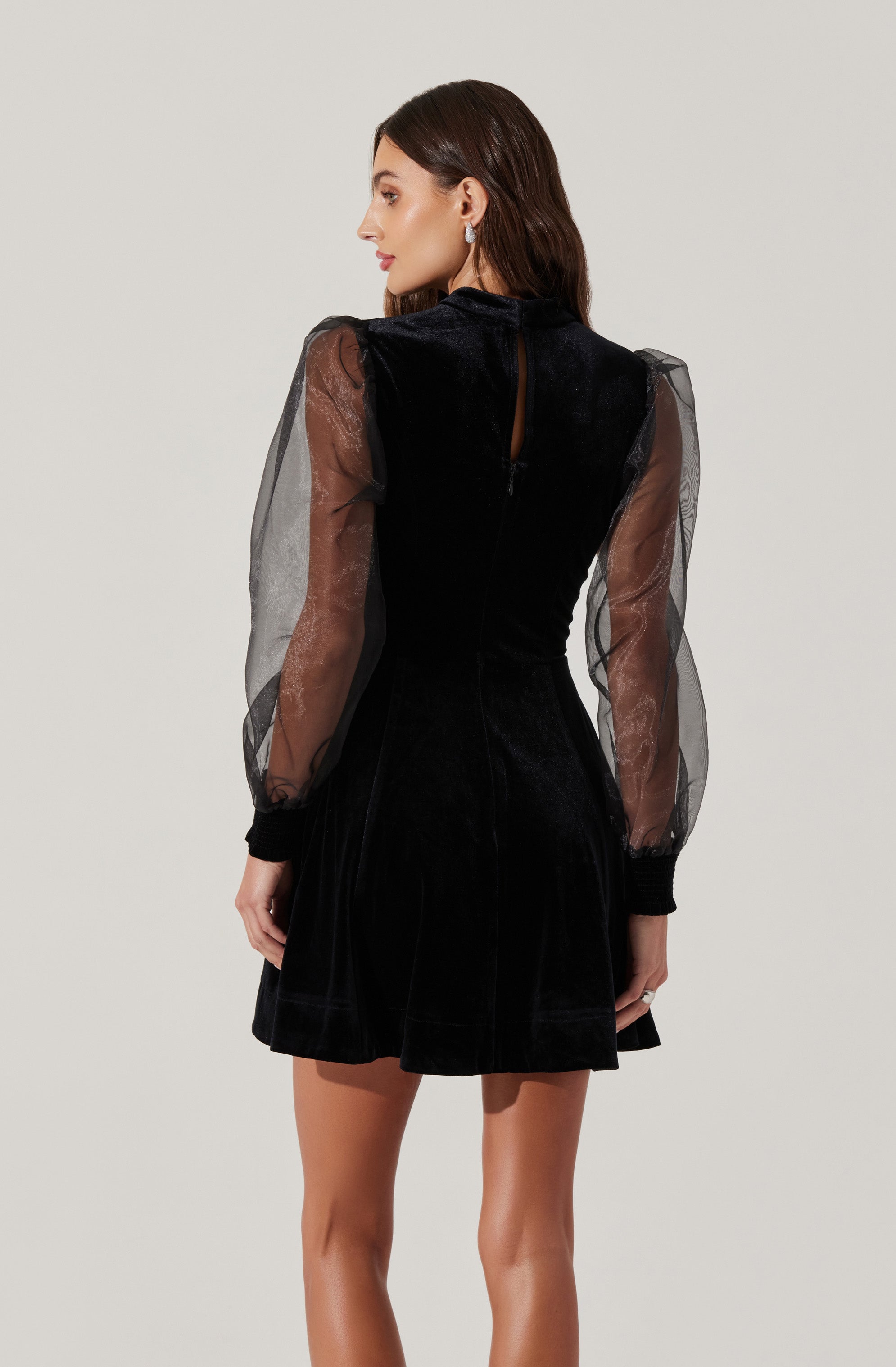 A woman is wearing a black dress with a velvet bodice and sheer, puffed long sleeves made of transparent fabric. The dress has a high neckline and a keyhole opening at the back with a button closure. The skirt of the dress is short and slightly flared.