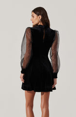 A woman is wearing a black dress with a velvet bodice and sheer, puffed long sleeves made of transparent fabric. The dress has a high neckline and a keyhole opening at the back with a button closure. The skirt of the dress is short and slightly flared.