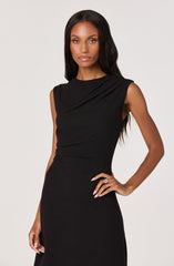 A woman is wearing a sleeveless black dress with a high neckline and subtle pleats on the shoulders. The dress is fitted at the waist and has a smooth, elegant silhouette. She has long, wavy black hair and is accessorizing with rings on her fingers.