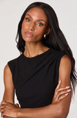 A woman is wearing a sleeveless black top with a high neckline and subtle pleats at the shoulder. She has long, dark hair and is accessorized with pearl earrings and a ring on her right hand, which is resting across her body.