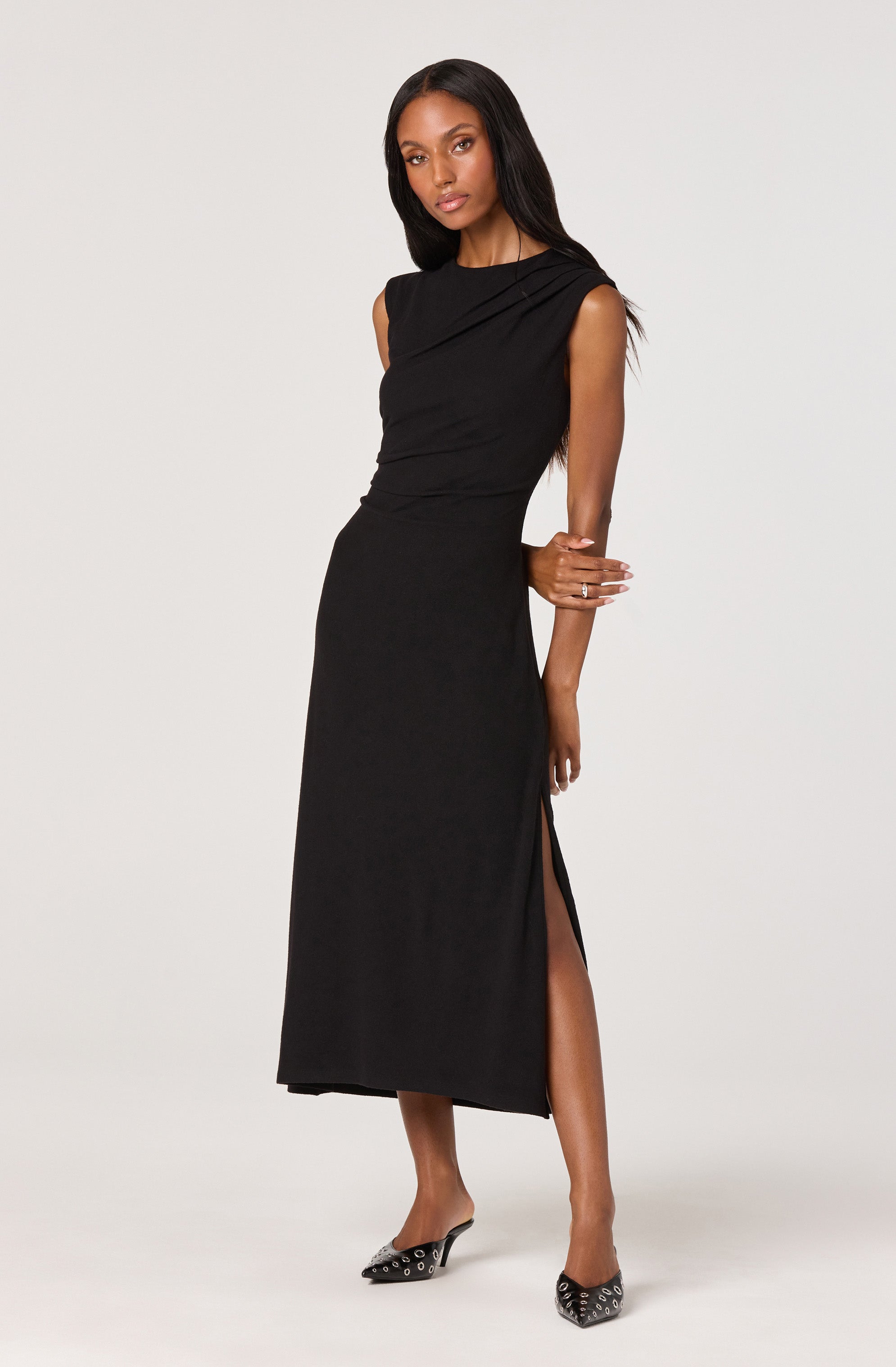 A woman is wearing a long, sleeveless black dress with a draped neckline and a high slit on one side. She has on black pointed-toe heels with metallic embellishments.