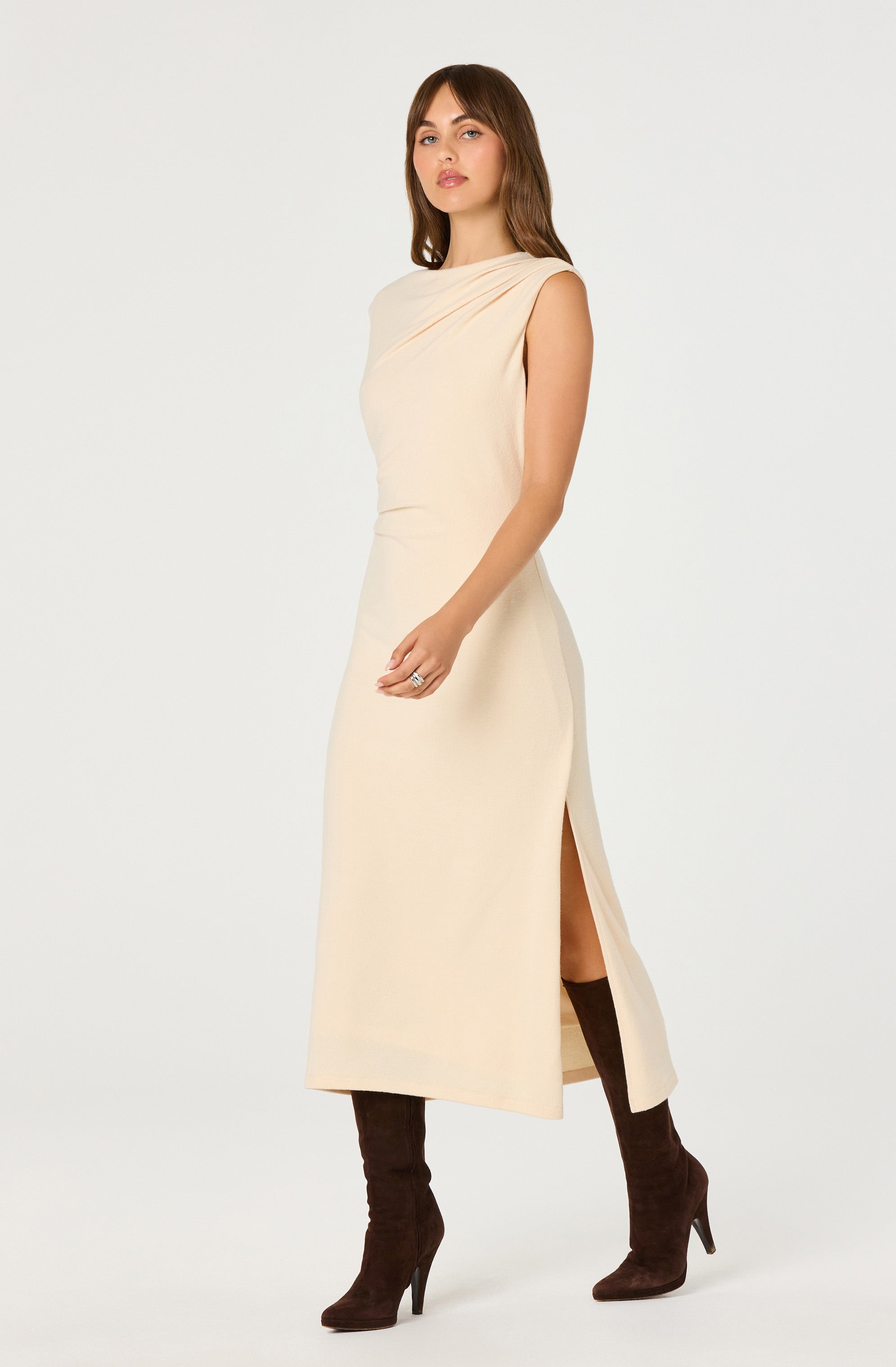 A woman is wearing a sleeveless, knee-length, creamy beige dress with a draped neckline and a side slit. She has on dark brown high-heeled boots.