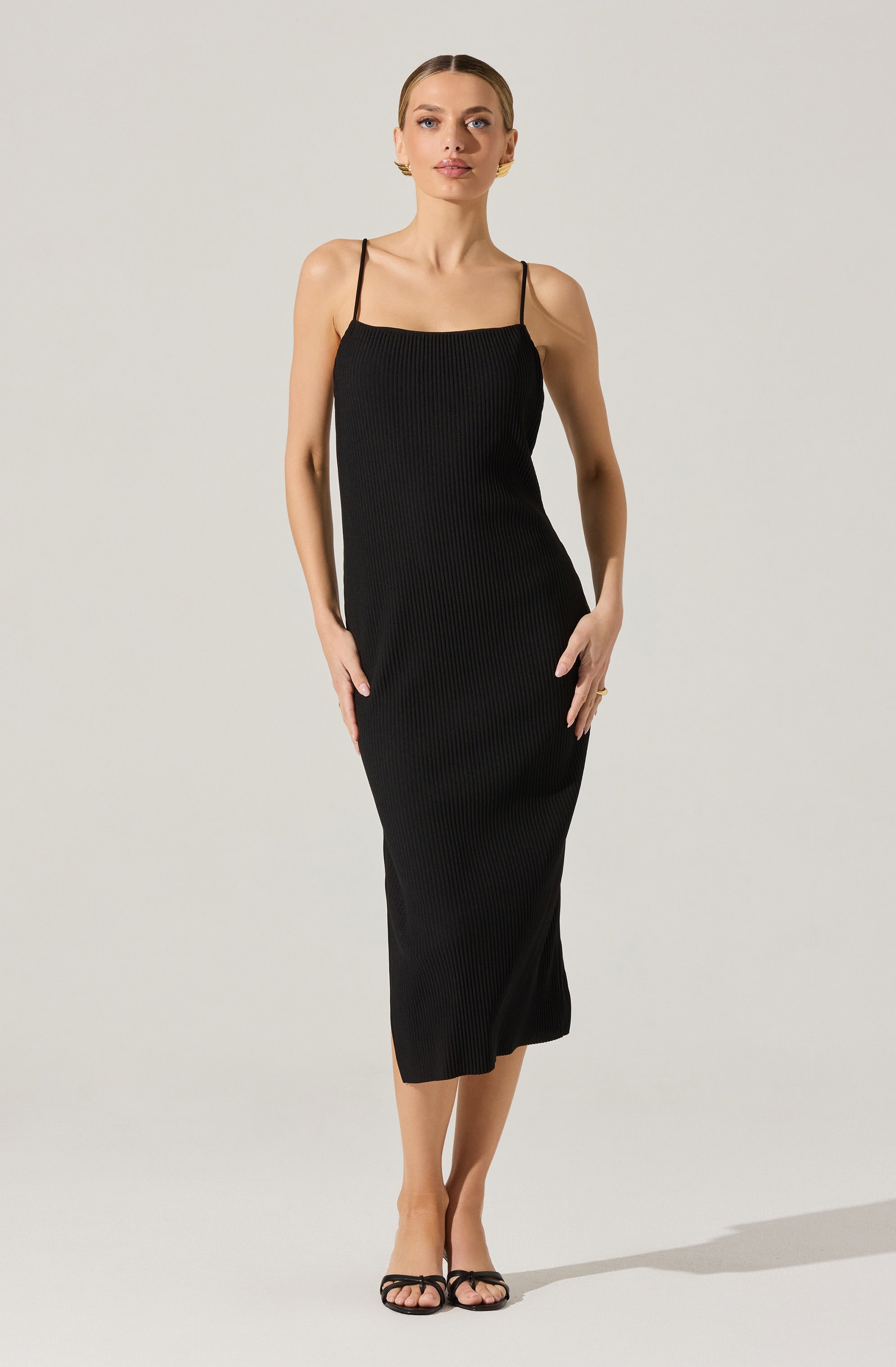 A woman is wearing a sleeveless black dress with thin spaghetti straps. The dress has a ribbed texture and falls to mid-calf length. She is wearing black open-toe sandals with thin straps.