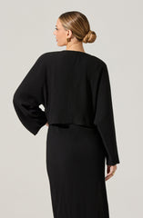 A woman is wearing a black, ribbed, long-sleeve top with a loose fit and wide sleeves. She pairs it with a matching black ribbed skirt that extends to the floor. Her hair is styled in a neat, low bun, and she is wearing gold earrings.