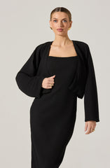 A woman is wearing a black, ribbed, fitted dress with a square neckline. She has a matching black, ribbed, cropped blazer draped over her shoulders.