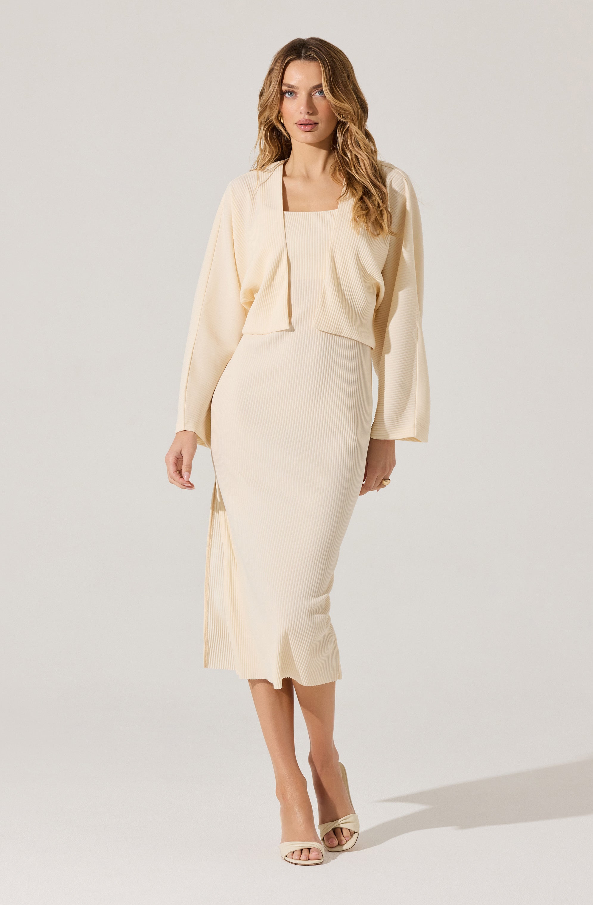 A woman is wearing a matching set in a light cream color. The outfit consists of a sleeveless, ribbed, square-neck top and a high-waisted, ribbed, midi-length skirt with a side slit. She has a loose, open-front cardigan draped over her shoulders with long sleeves. She is also wearing light-colored, open-toe high heels.