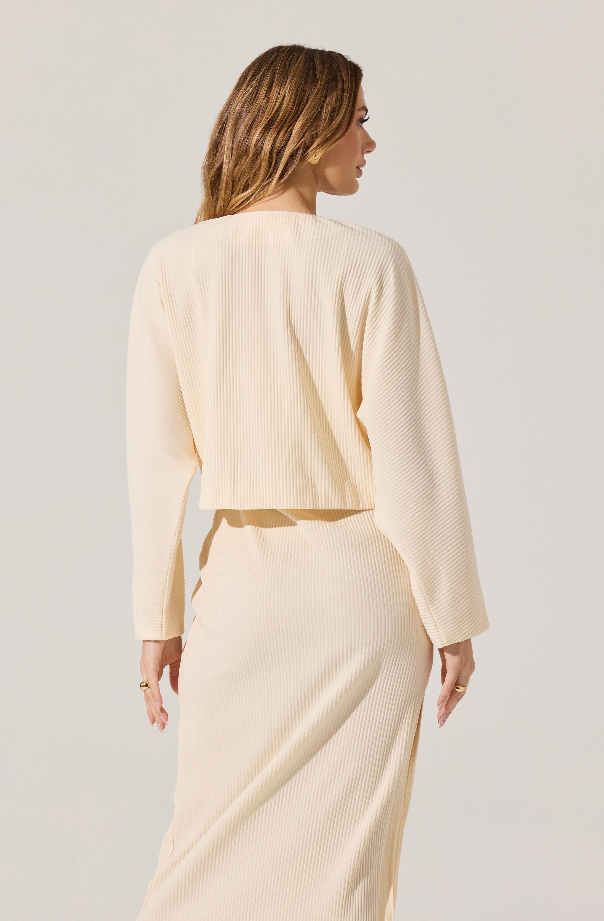 A woman is wearing a cream-colored, ribbed knit two-piece outfit consisting of a long-sleeved, cropped top and a matching high-waisted skirt. The fabric has a vertical ribbed texture, and the top has a relaxed fit with a round neckline. The skirt extends down to her ankles, creating a coordinated and elegant look.