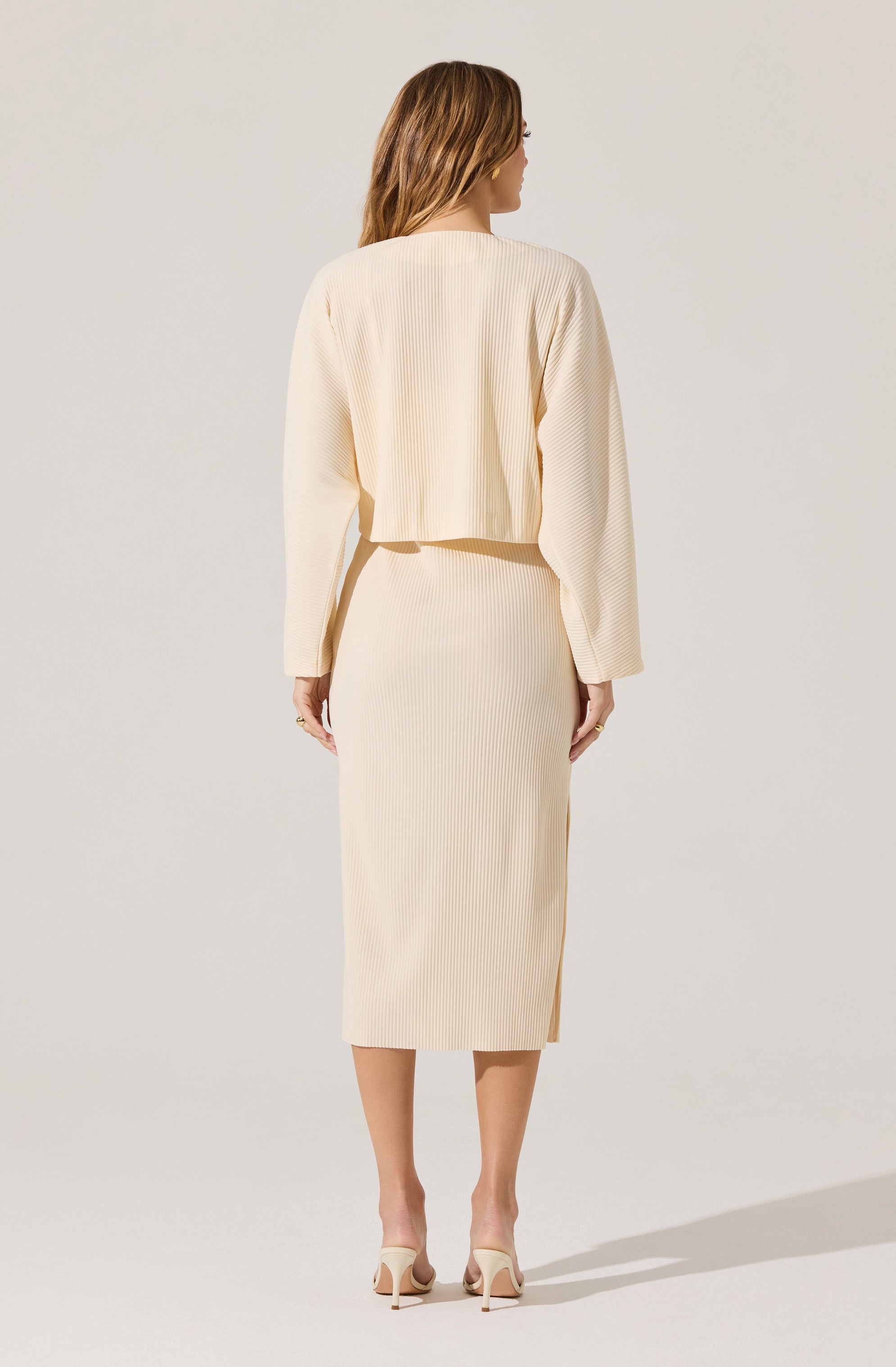 A woman is wearing a matching cream-colored outfit consisting of a cropped, long-sleeved, textured top and a high-waisted, ribbed midi skirt. She is also wearing light-colored, open-toe high heels. The overall style appears elegant and minimalistic.