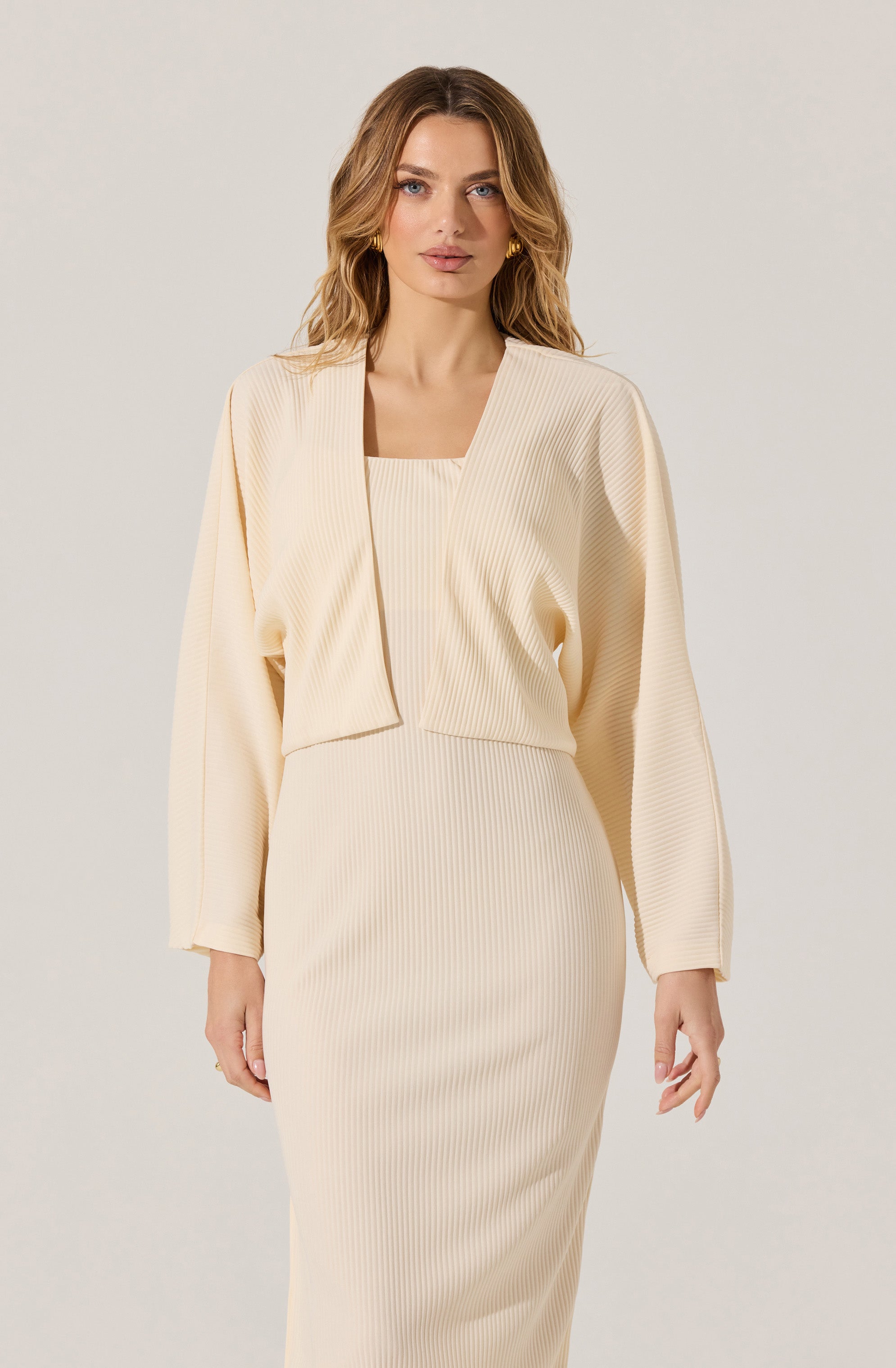A woman is wearing a cream-colored, ribbed knit outfit consisting of a fitted, high-waisted skirt and a matching cropped jacket with long sleeves. The jacket has a relaxed fit with a draped, open front.