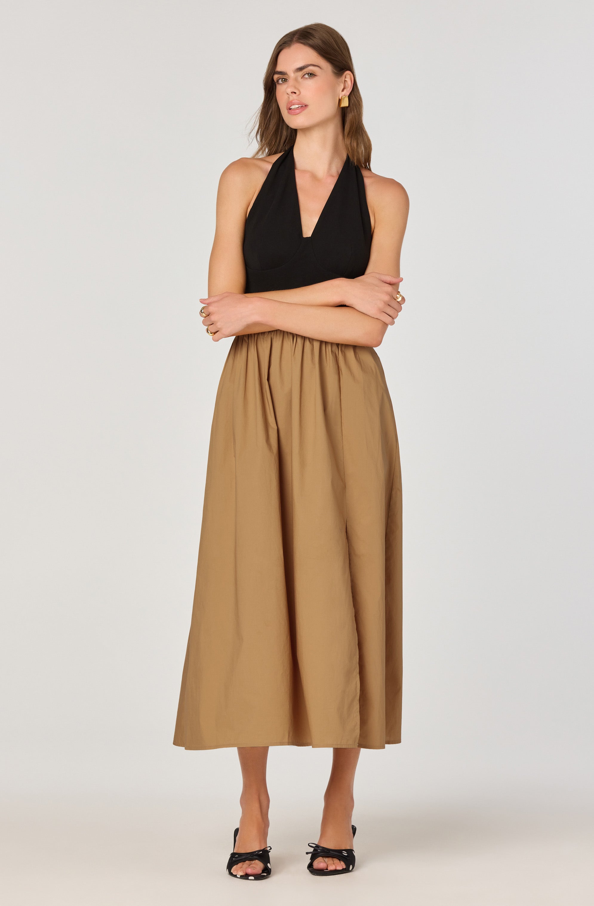 A woman is wearing a sleeveless black top with a V-neckline and wide shoulder straps. She pairs it with a high-waisted, full-length beige skirt that has an elastic waistband. She completes her outfit with black open-toed high-heeled sandals. She has long brown hair, gold earrings, and a relaxed pose with arms crossed.