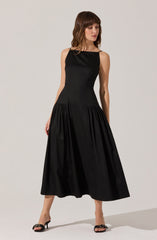 A woman is wearing a sleeveless, ankle-length black dress with a fitted bodice and a full, gathered skirt. She has on black high-heeled sandals with decorative bows on the front.