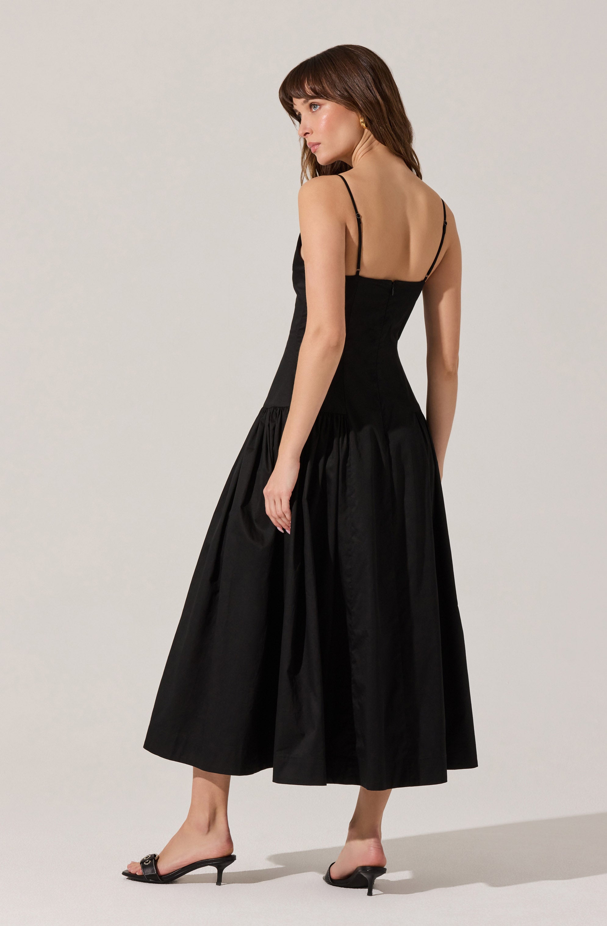 A woman is wearing a black dress with thin spaghetti straps, a fitted bodice, and a high waist. The skirt is long, full, and gathered, creating a voluminous and elegant appearance. She is also wearing black open-toe high-heeled sandals.