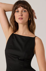 A woman is wearing a sleeveless black dress with a boat neckline. The dress appears to be made of a smooth, shiny fabric that fits closely to her body. She accessorized with small, gold hoop earrings.
