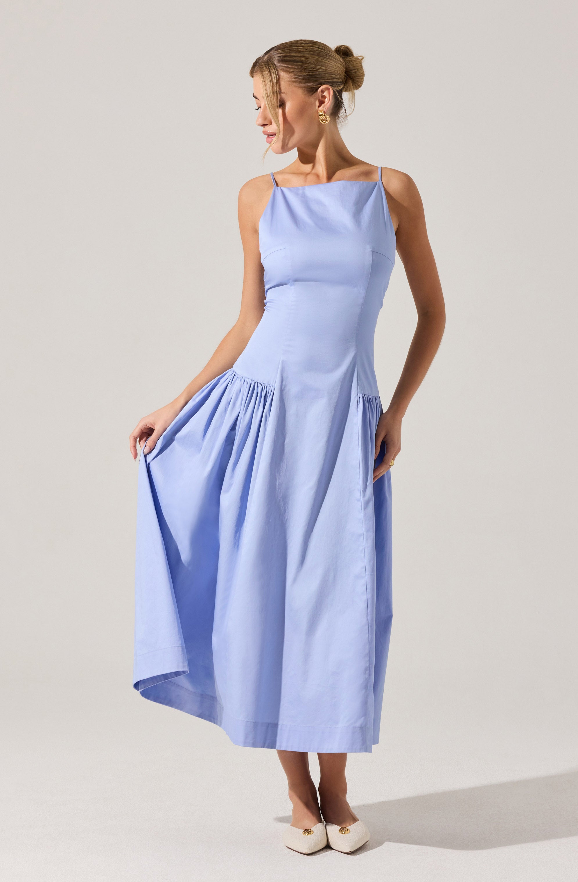 A woman is wearing a light blue sleeveless dress with thin spaghetti straps. The dress has a fitted bodice and a gathered skirt that she is holding up slightly with her left hand. She is also wearing cream-colored shoes with embellishments at the front. Her hair is styled in a neat, twisted bun.