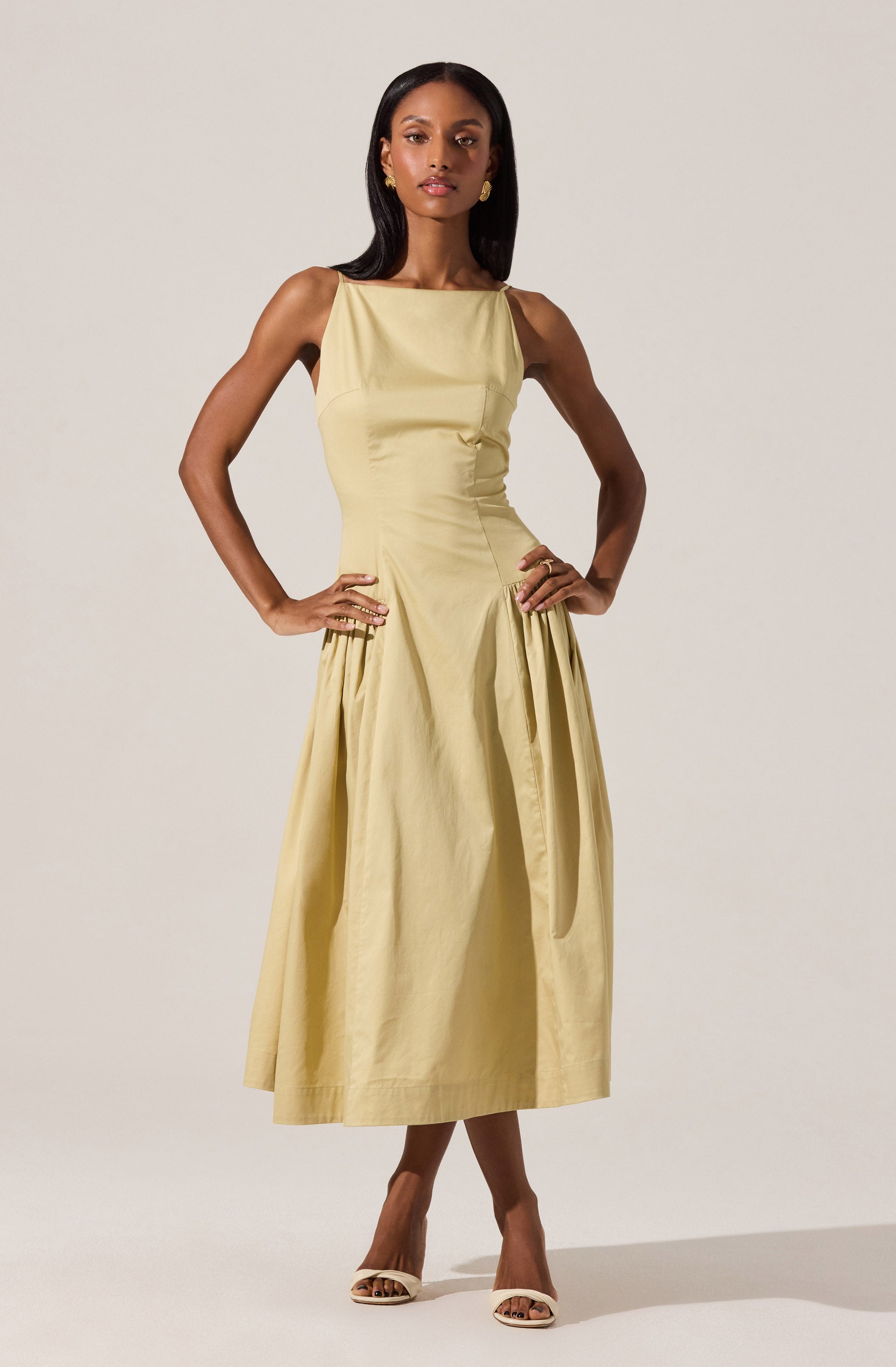A woman is wearing a sleeveless, light yellow midi dress with a fitted waist and a full, pleated skirt. The dress has a smooth, shiny fabric with a simple, elegant design, and she pairs it with white high-heeled shoes.