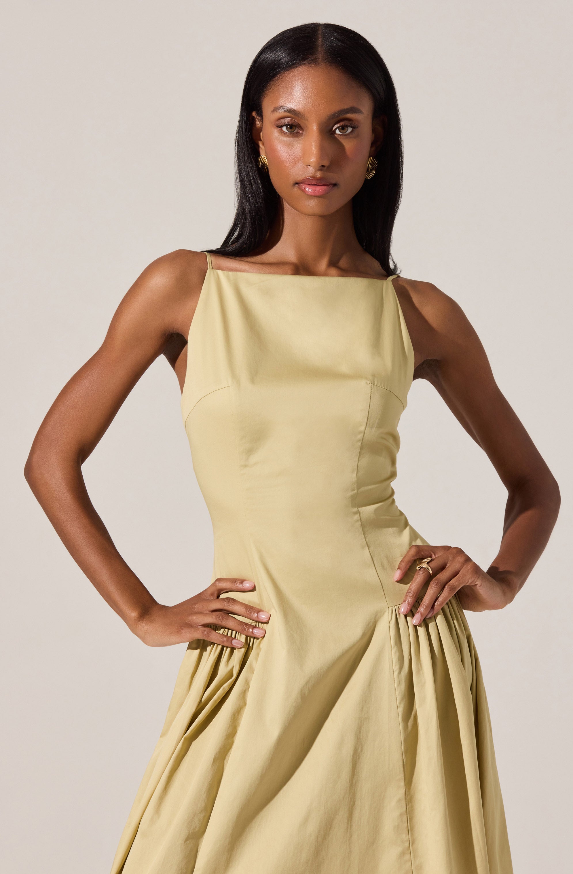 A woman is wearing a sleeveless, floor-length, satin-like yellow gown with a high, square neckline and structured bodice. The dress features gathered detailing at the waist and around the hips, creating a voluminous skirt. The material has a smooth, shiny texture, and she accessorizes with gold earrings and a gold ring.