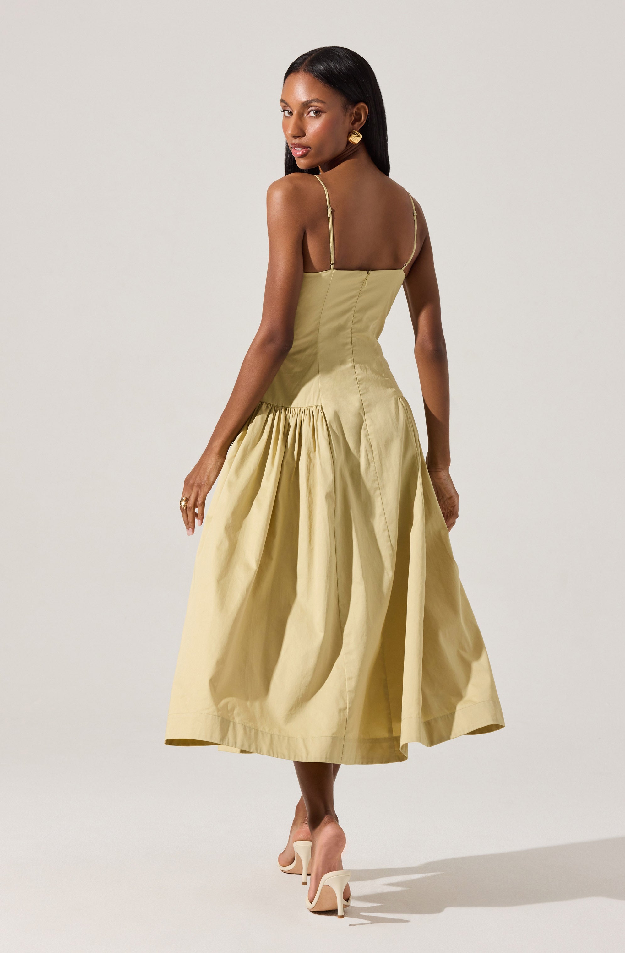 A woman is wearing a light yellow, sleeveless dress with thin spaghetti straps. The dress features a fitted bodice and a full, gathered skirt that falls to mid-calf length. She is also wearing light-colored high-heeled shoes.