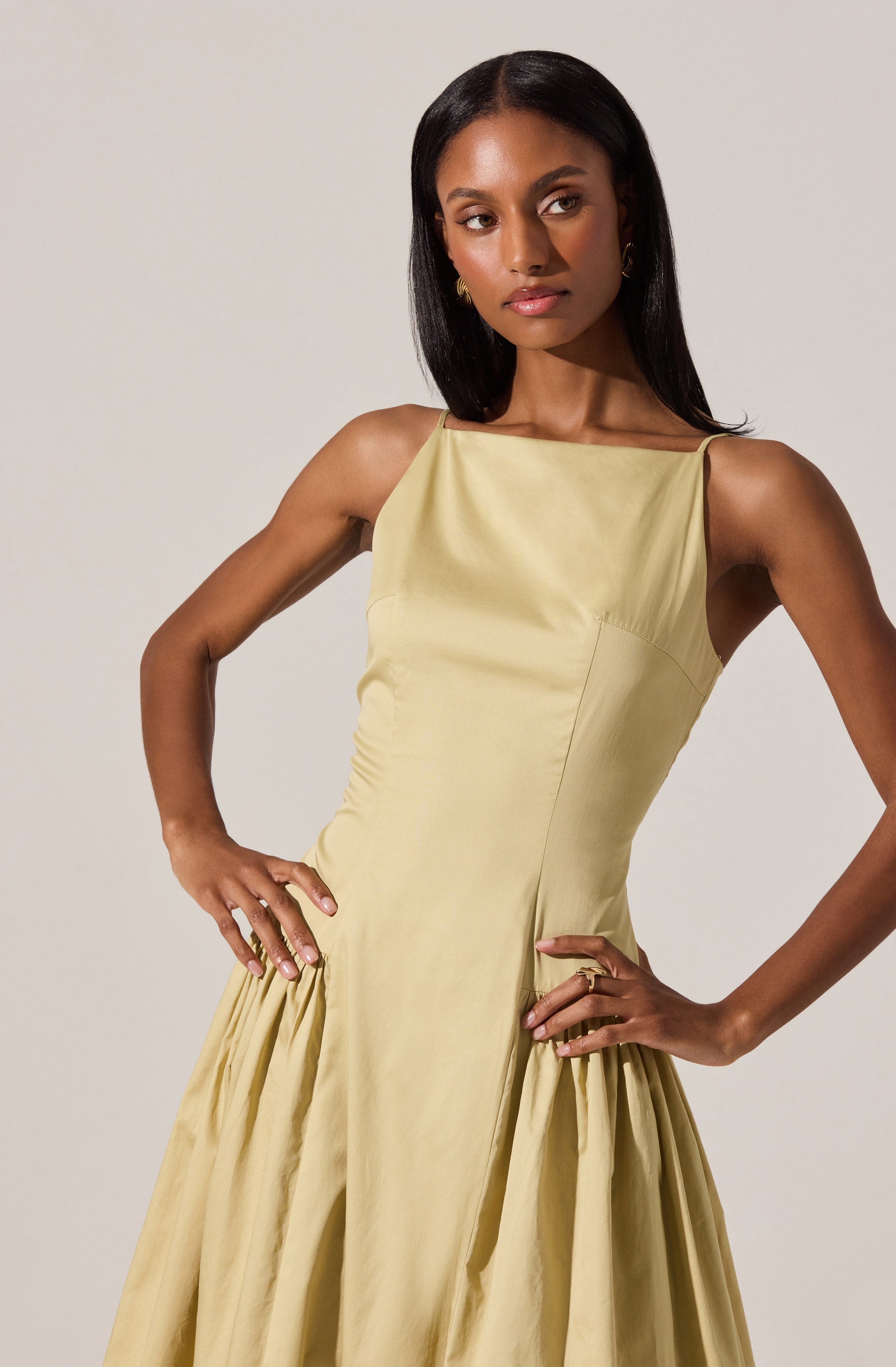A woman is wearing a sleeveless, light yellow dress with a fitted bodice and a full skirt. The dress features a bateau neckline and gathered fabric at the waistline, creating volume in the skirt.