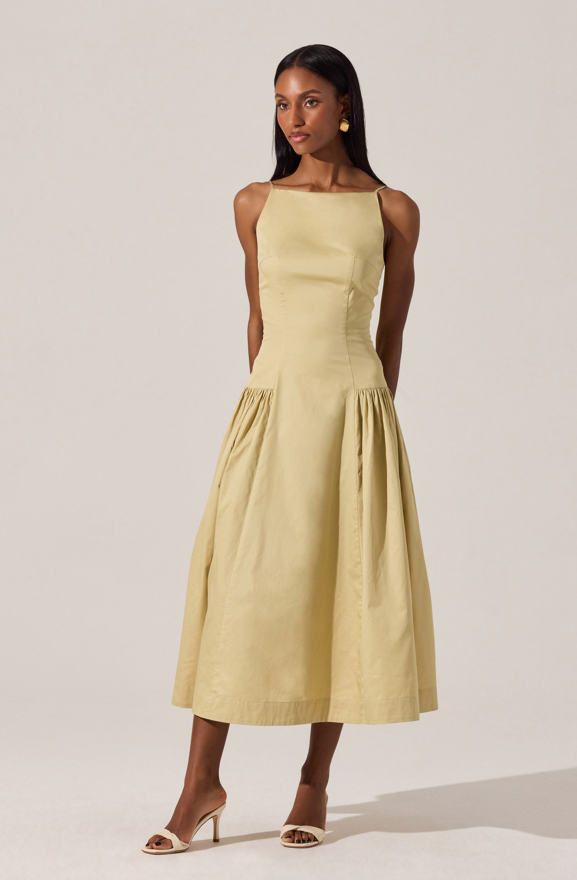 A woman is wearing a sleeveless, ankle-length, pale yellow dress with a fitted bodice and a full skirt that has gathered fabric at the hips. She is accessorizing with gold earrings and is wearing beige high-heeled sandals.