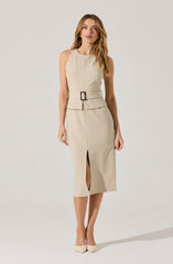A woman is wearing a sleeveless, beige midi dress with a fitted waist accented by a wide belt with a large rectangular buckle. The dress features a front slit at the hem, adding a stylish detail. She is also wearing high-heeled shoes that match the color of the dress.