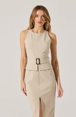 A woman is wearing a sleeveless, beige dress with a fitted bodice and a round neckline. The dress features a wide belt at the waist with a rectangular black and beige buckle. The skirt of the dress has a front slit.