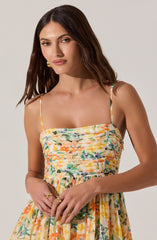 The woman is wearing a sleeveless dress with thin spaghetti straps. The top part of the dress is fitted and ruched, featuring a colorful floral print in shades of yellow, orange, green, purple, and blue. The skirt is gathered at the waist with a similar vibrant floral pattern, creating a flowy silhouette.