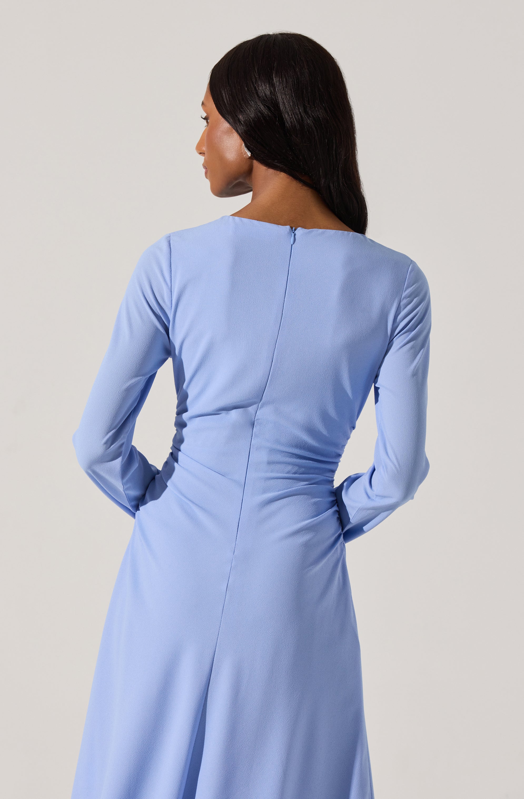 A woman is wearing a long, light blue dress with long sleeves and a round neckline. The dress has a fitted waist and appears to be made of a smooth fabric with a slight sheen. The back of the dress features a central seam running from the neckline to the waist, with a hidden zipper closure.