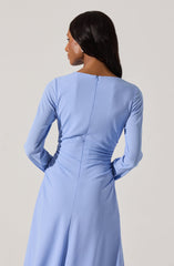 A woman is wearing a long, light blue dress with long sleeves and a round neckline. The dress has a fitted waist and appears to be made of a smooth fabric with a slight sheen. The back of the dress features a central seam running from the neckline to the waist, with a hidden zipper closure.